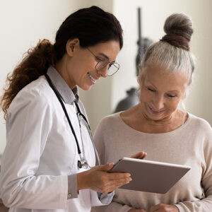 Happy female doctor and senior patient look at tablet screen discuss treatment or therapy on gadget. Smiling woman nurse and mature client use pad device discussing results in modern hospital.