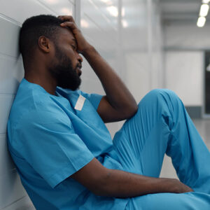 Stressed tired young afro surgeon sitting on floor in hospital corridor after difficult surgery. Side view of afro-american doctor in scrubs resting on hallway floor feeling exhausted