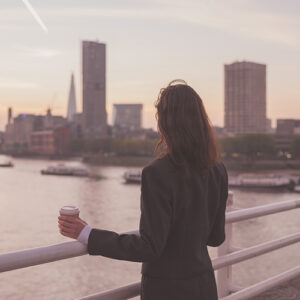 A young businesswoman is standing on a bridge with a cup of coffee in her hand and is admiring the sun rise over the London skyline